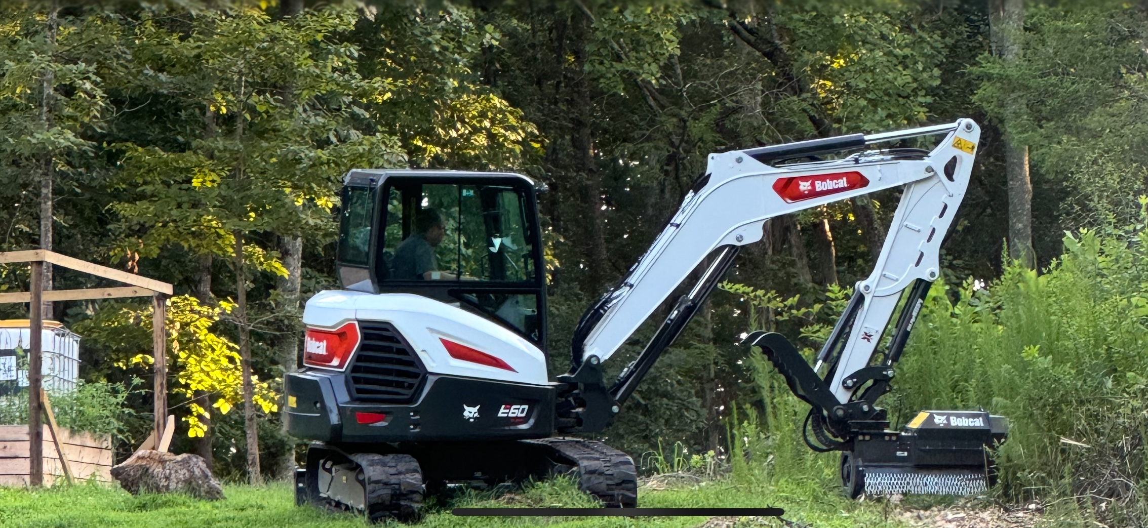 A white and black Bobcat E60 compact excavator with a brush cutter attachment is parked on grass near a wooded area.