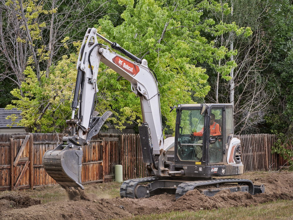 A person operates a Bobcat excavator, digging earth in a fenced yard with trees in the background.