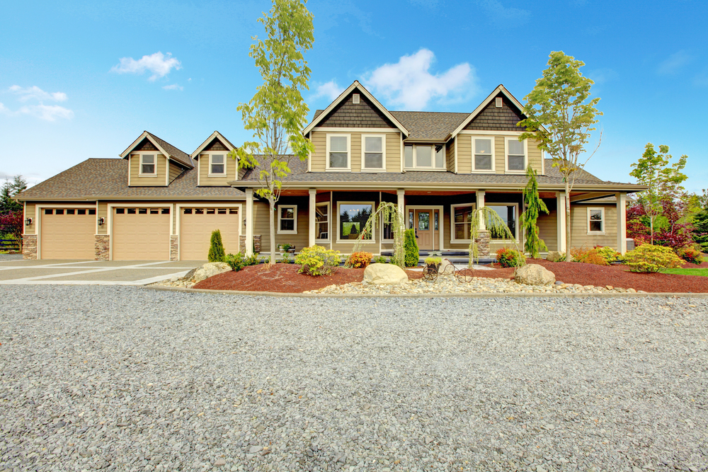 Large two-story suburban house with three-car garage, front porch, landscaped yard, gravel driveway, and clear blue sky.