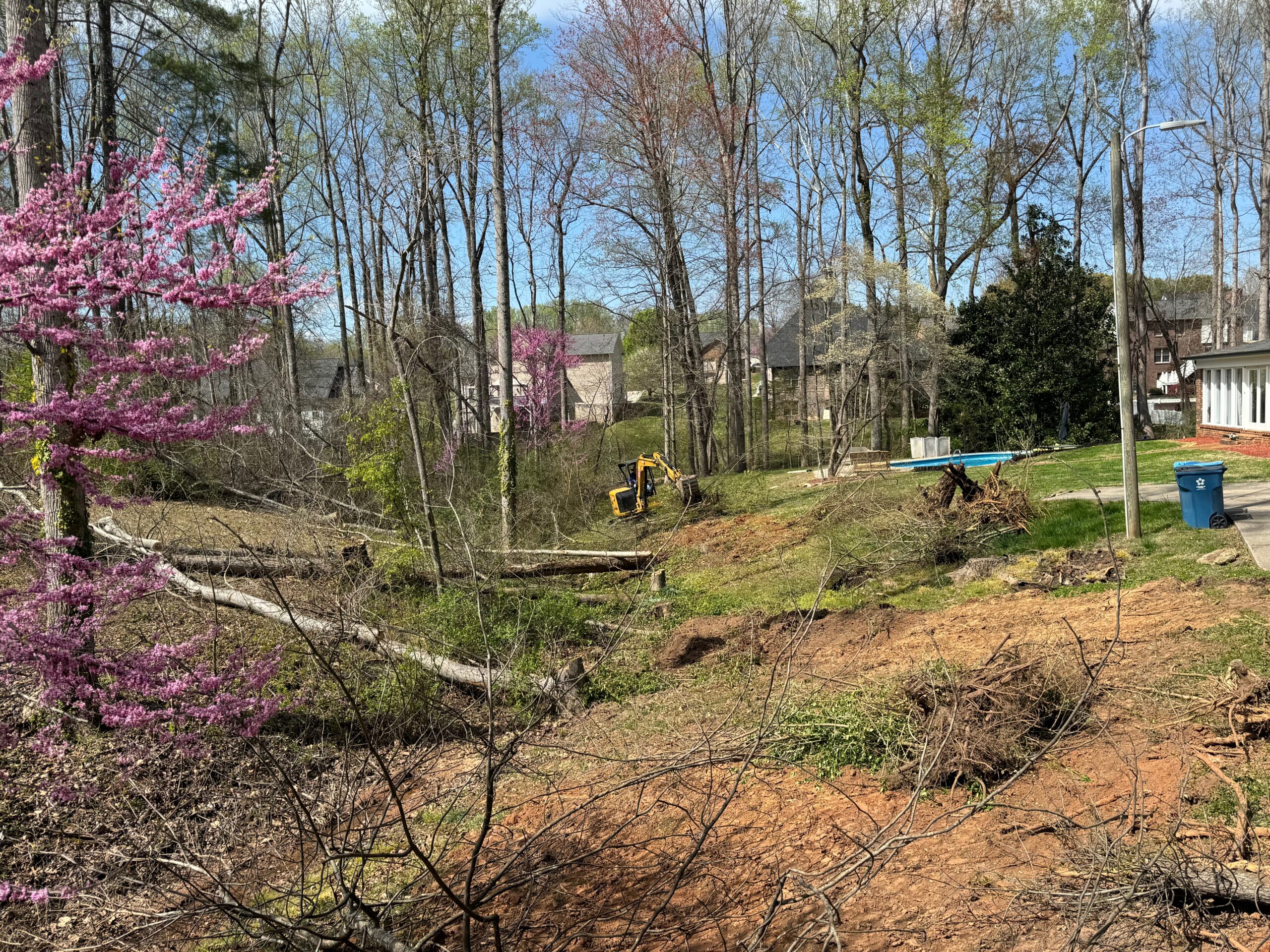 A small yellow excavator works on clearing fallen trees and brush in a wooded residential area with visible houses and a blue trash bin.